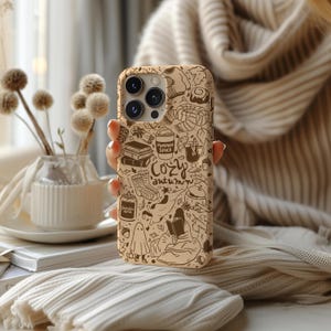 May include: A tan phone case with a cosy autumn design. The case features illustrations of books, coffee, a ghost, and the words "Pumpkin Spice" and "Cozy Autumn". The case is held in a person's hand.