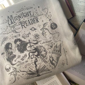 May include: A beige sweatshirt featuring a black ink-style graphic with the text "MIDNIGHT READER", "*plot twist*", and "DEATH BY TBR". The design includes illustrations of a castle, a skull, a potion bottle, a sword, a rose, and a book.