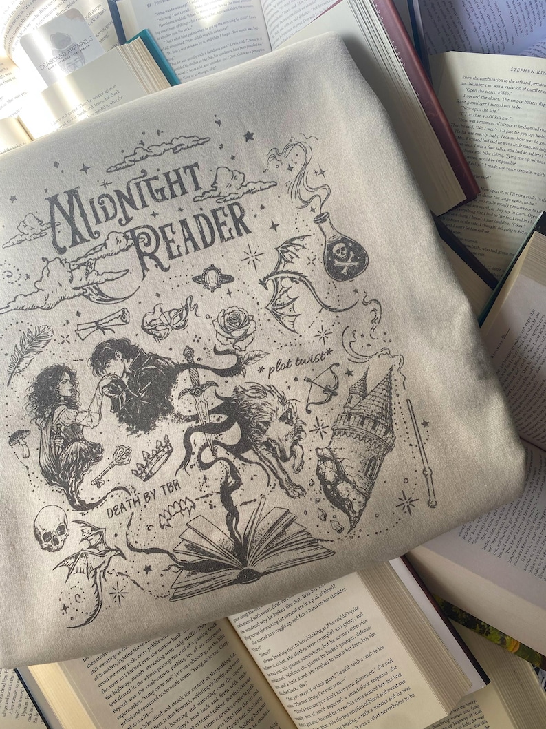 Op de afbeelding: Een beige sweatshirt met een zwarte inktstijl grafische print met de tekst "MIDNIGHT READER", "*plot twist*" en "DEATH BY TBR". Het ontwerp bevat illustraties van een kasteel, een schedel, een toverdrankflesje, een zwaard, een roos en een boek.