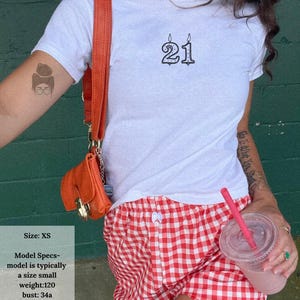 May include: White t-shirt with a black '21' candle design. The model is wearing red and white checkered shorts and carrying an orange purse. A pink drink with a straw is also visible. The model's size is XS.