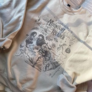 Op de afbeelding: Een beige sweater met ronde hals en een donkergrijze grafische print met de tekst "MIDNIGHT READER", "DEATH BY TBR" en "plot twist". Het ontwerp bevat illustraties van een koppel, een schedel, een kroon, een zwaard, een toverdrankfles en een boek.