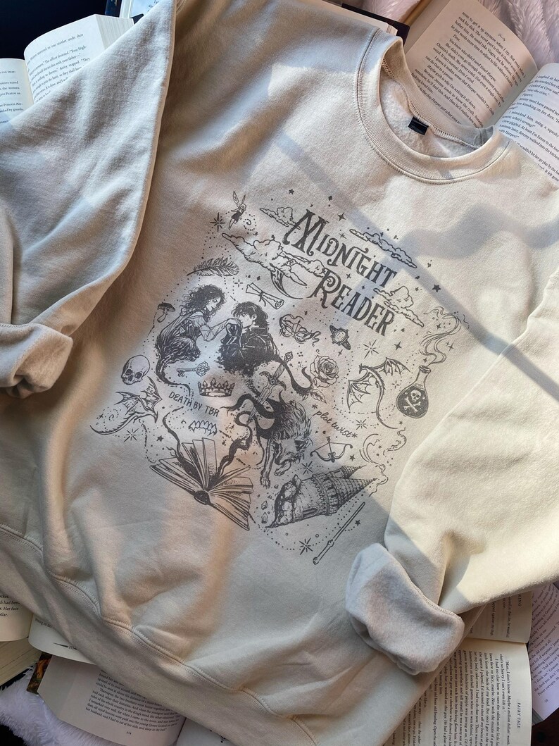 Op de afbeelding: Een beige sweater met ronde hals en een donkergrijze grafische print met de tekst "MIDNIGHT READER", "DEATH BY TBR" en "plot twist". Het ontwerp bevat illustraties van een koppel, een schedel, een kroon, een zwaard, een toverdrankfles en een boek.