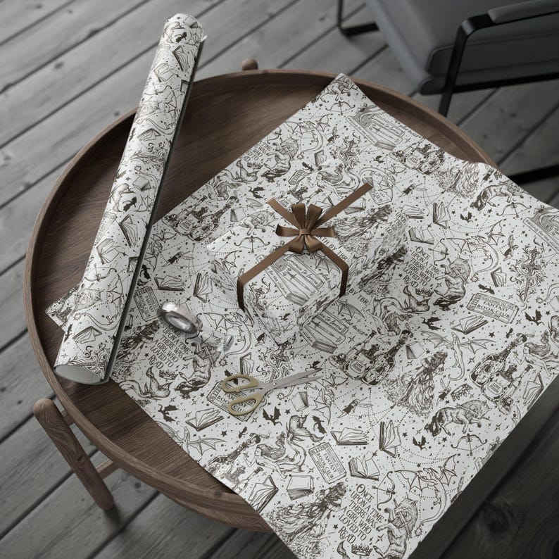 May include: A roll of white wrapping paper and a wrapped gift with a brown ribbon, both featuring a black and white fantasy illustration. A pair of gold scissors and tape are also on the wooden table. The wrapping paper has text that reads "Once upon a time there was a girl who loved to read."
