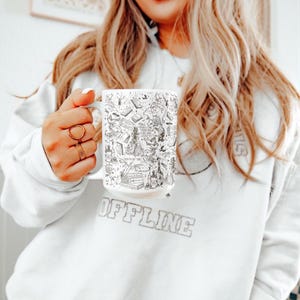 K&ouml;nnte beinhalten: Eine wei&szlig;e Keramiktasse mit einer schwarz-wei&szlig;en Illustration zu einem Buchthema. Das Design enth&auml;lt verschiedene Buchtitel, Figuren und Objekte, die mit B&uuml;chern und Lesen zu tun haben. Die Tasse wird von einer Person gehalten, die ein wei&szlig;es Sweatshirt mit dem Aufdruck "OFFLINE" tr&auml;gt.