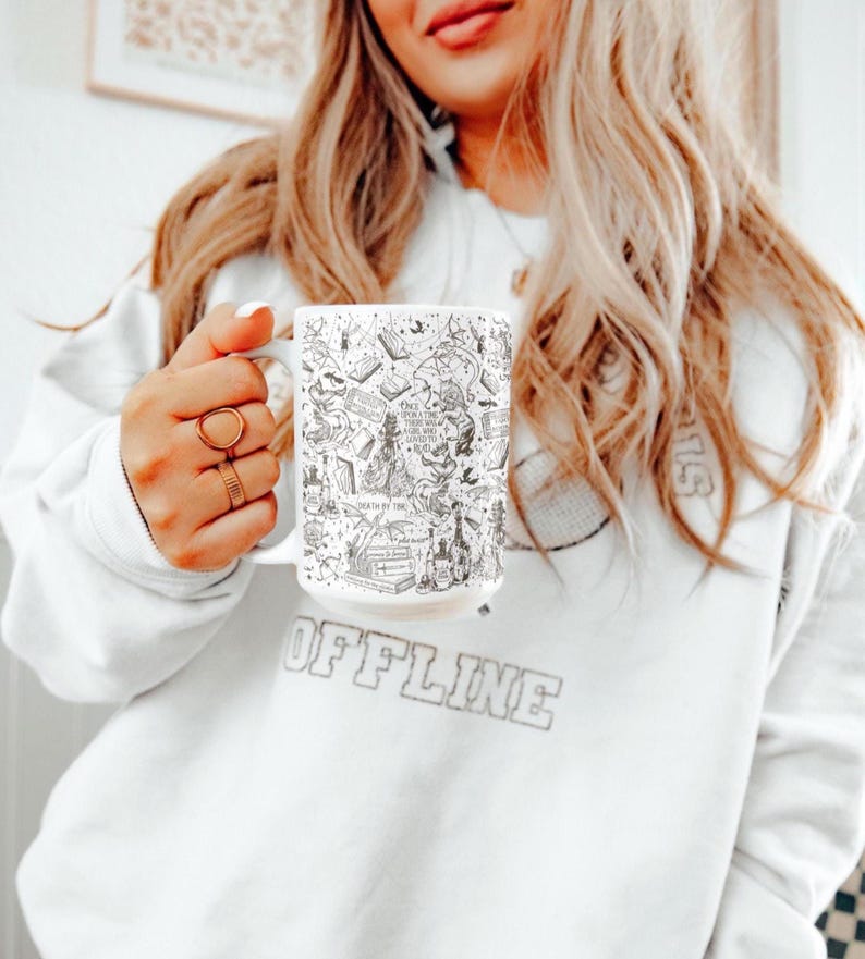 K&ouml;nnte beinhalten: Eine wei&szlig;e Keramiktasse mit einer schwarz-wei&szlig;en Illustration zu einem Buchthema. Das Design enth&auml;lt verschiedene Buchtitel, Figuren und Objekte, die mit B&uuml;chern und Lesen zu tun haben. Die Tasse wird von einer Person gehalten, die ein wei&szlig;es Sweatshirt mit dem Aufdruck "OFFLINE" tr&auml;gt.