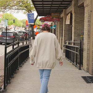 Op de afbeelding: Een oversized beige sweater met ronde hals, gedragen met een lichtblauwe spijkerbroek, een bordeauxrode honkbalpet en witte sneakers. De sweater heeft een losse pasvorm met lange mouwen en een geribde zoom voor een neutrale uitstraling.