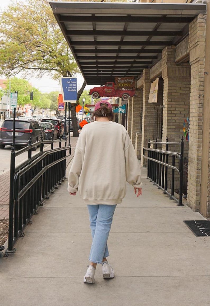 Op de afbeelding: Een oversized beige sweater met ronde hals, gedragen met een lichtblauwe spijkerbroek, een bordeauxrode honkbalpet en witte sneakers. De sweater heeft een losse pasvorm met lange mouwen en een geribde zoom voor een neutrale uitstraling.