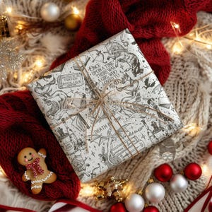 May include: A wrapped gift with a fantasy book theme, featuring black and white illustrations and text. The wrapping paper is tied with twine and rests on a red knitted scarf and a cream-colored blanket, with Christmas ornaments and a gingerbread man.