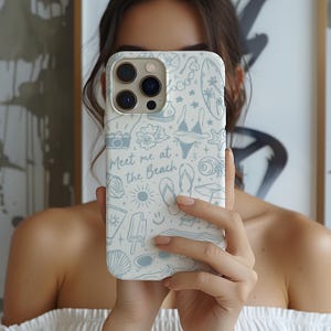 K&ouml;nnte beinhalten: Eine wei&szlig;e Handyh&uuml;lle mit einem hellblauen Strandmuster. Das Design umfasst Illustrationen von Muscheln, einem Bikini, einem Surfbrett und dem Text "Meet me at the Beach". Die H&uuml;lle wird vor einer Person gehalten.