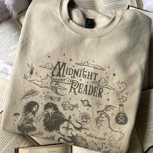 Op de afbeelding: Een beige sweater met ronde hals en donkerbruine zeefdruk. Het ontwerp bevat de tekst "Midnight Reader" en "plot twist", omringd door illustraties van een koppel, een zwaard, een roos, een toverdrankflesje en sterren.