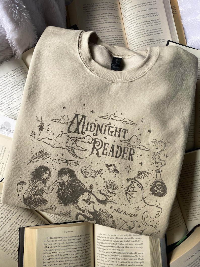 Op de afbeelding: Een beige sweater met ronde hals en donkerbruine zeefdruk. Het ontwerp bevat de tekst "Midnight Reader" en "plot twist", omringd door illustraties van een koppel, een zwaard, een roos, een toverdrankflesje en sterren.