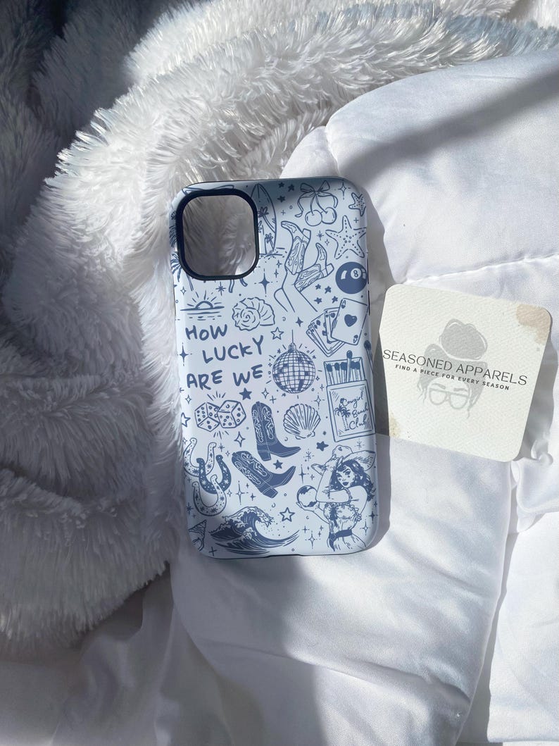 Puede incluir: Funda de tel&eacute;fono azul claro con un patr&oacute;n repetido de varias ilustraciones, incluyendo una bola de discoteca, botas de vaquero y naipes. La frase "HOW LUCKY ARE WE" est&aacute; escrita en la funda. Una peque&ntilde;a tarjeta con texto est&aacute; al lado.