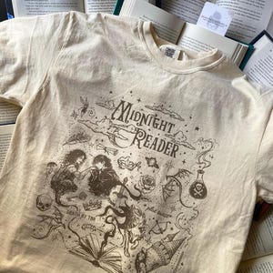 Puede incluir: Una camiseta de color crema con un estampado gr&aacute;fico marr&oacute;n oscuro con el texto "Midnight Reader", "Death by TBR" y "plot twist". La ilustraci&oacute;n incluye una pareja, una calavera, una rosa, una poci&oacute;n, una corona, un lobo y un castillo.