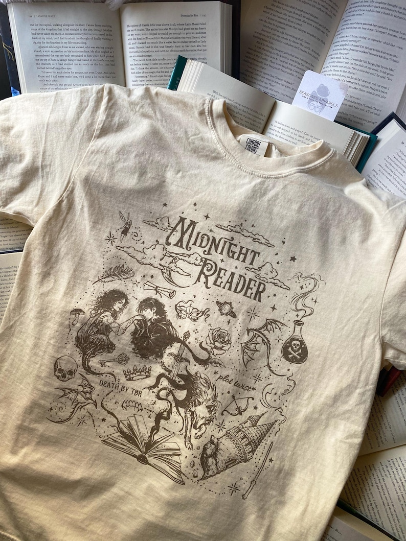 Puede incluir: Una camiseta de color crema con un estampado gr&aacute;fico marr&oacute;n oscuro con el texto "Midnight Reader", "Death by TBR" y "plot twist". La ilustraci&oacute;n incluye una pareja, una calavera, una rosa, una poci&oacute;n, una corona, un lobo y un castillo.