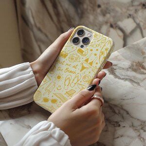 May include: A white iPhone case with a yellow design featuring a variety of summer-themed illustrations, including flip flops, sunglasses, a seashell, and the text "Meet me at the beach".