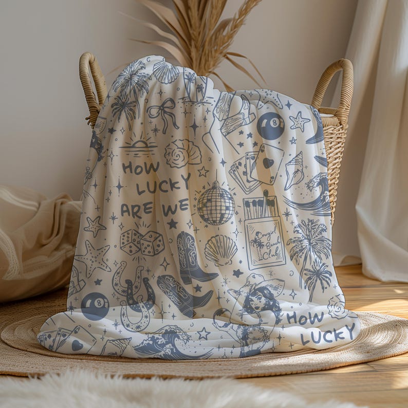 May include: A white throw blanket with a blue and white pattern of various lucky charms, including horseshoes, dice, stars, palm trees, seashells, and a disco ball. The text "How lucky are we" is repeated on the blanket.