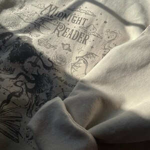 Op de afbeelding: Een cr&egrave;mekleurig sweatshirt met ronde hals en een donkergrijze grafische print met de tekst "MIDNIGHT READER" en "plot twist". Het ontwerp bevat illustraties van een draak, een zwaard, een roos, sterren, wolken en een open boek.
