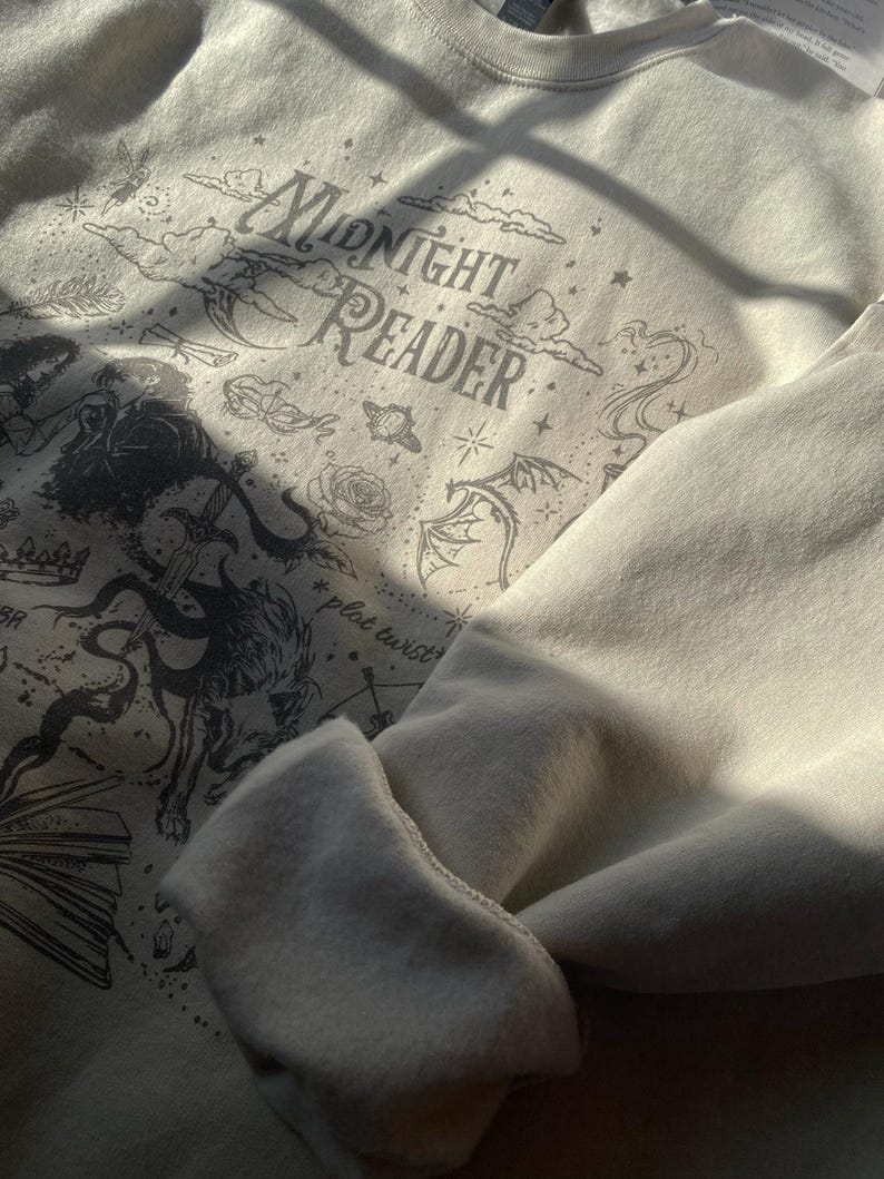 Op de afbeelding: Een cr&egrave;mekleurig sweatshirt met ronde hals en een donkergrijze grafische print met de tekst "MIDNIGHT READER" en "plot twist". Het ontwerp bevat illustraties van een draak, een zwaard, een roos, sterren, wolken en een open boek.