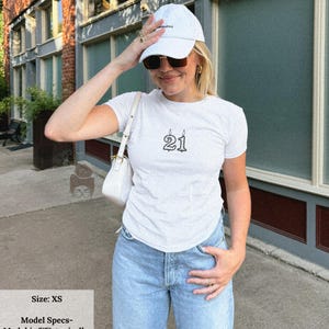 May include: White t-shirt with the number "21" printed in black, styled with light blue jeans and a white baseball cap. The model is wearing an XS for a baby tee fit. The image is taken outdoors.