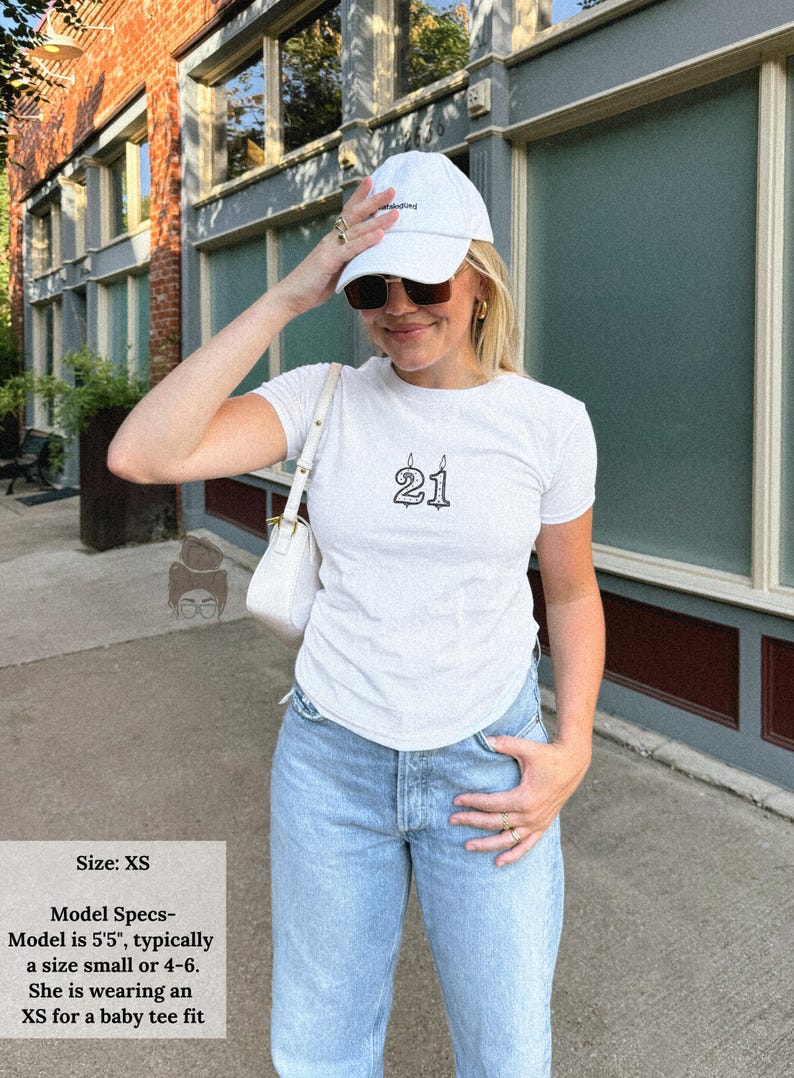 May include: White t-shirt with the number "21" printed in black, styled with light blue jeans and a white baseball cap. The model is wearing an XS for a baby tee fit. The image is taken outdoors.