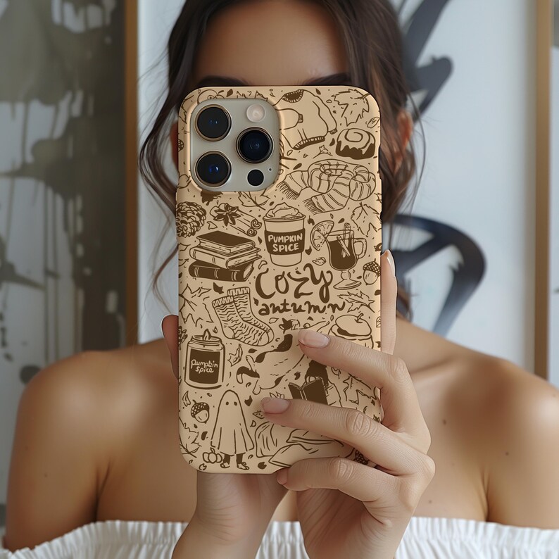 May include: A beige phone case featuring a repeating pattern of autumn-themed illustrations, including a coffee cup, books, a cosy sock, and the words "Cozy Autumn" in a handwritten font. The case is held up in front of a person.