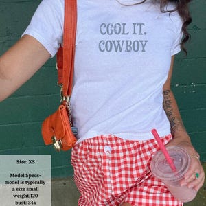 May include: White t-shirt with the text "COOL IT, COWBOY" in gray, paired with red and white checkered shorts. An orange purse and a pink drink with a straw are also visible.