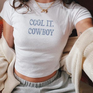 May include: White crop top with the text "COOL IT, COWBOY" in light blue. The shirt is short-sleeved and has a round neckline. The model is wearing gray sweatpants and a gold necklace.