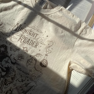 Puede incluir: Una camiseta de manga corta de color crema con un estampado gr&aacute;fico marr&oacute;n oscuro con el texto "Midnight Reader" y "plot twist". El dise&ntilde;o incluye ilustraciones de una botella de poci&oacute;n con una calavera, una rosa, un castillo y estrellas.