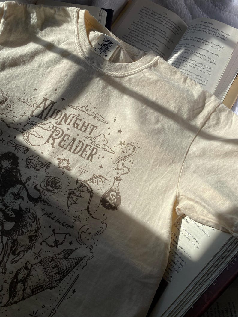 Puede incluir: Una camiseta de manga corta de color crema con un estampado gr&aacute;fico marr&oacute;n oscuro con el texto "Midnight Reader" y "plot twist". El dise&ntilde;o incluye ilustraciones de una botella de poci&oacute;n con una calavera, una rosa, un castillo y estrellas.
