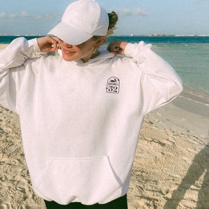 Puede incluir: Sudadera con capucha blanca con un gr&aacute;fico negro de una palmera y el texto "Isla 52" en la parte delantera. La persona lleva una gorra de b&eacute;isbol blanca.