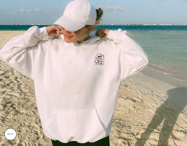 Puede incluir: Sudadera con capucha blanca con un gr&aacute;fico negro de una palmera y el texto "Isla 52" en la parte delantera. La persona lleva una gorra de b&eacute;isbol blanca.