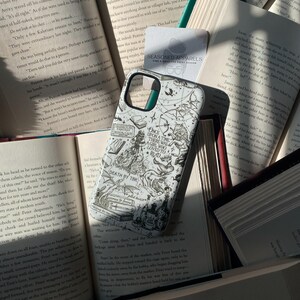 May include: A white smartphone case featuring black line art illustrations of books, a wolf, a dragon, and text that reads Once Upon a Time There Was a Girl Who Loved to Read and Death by TBR, resting on top of several open hardcover books.