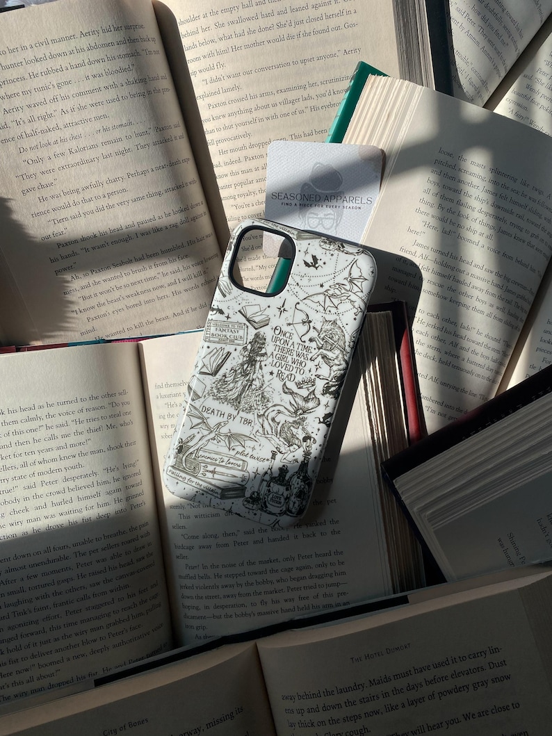 May include: A white smartphone case featuring black line art illustrations of books, a wolf, a dragon, and text that reads Once Upon a Time There Was a Girl Who Loved to Read and Death by TBR, resting on top of several open hardcover books.