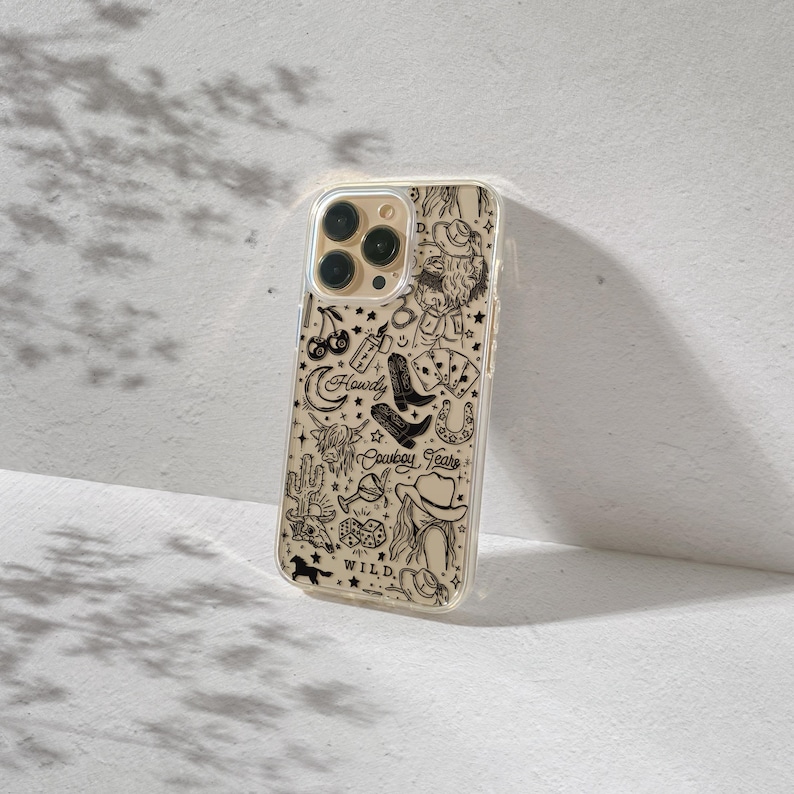 Puede incluir: Una funda transparente para tel&eacute;fono con un fondo beige y un dise&ntilde;o de tem&aacute;tica vaquera en negro. El dise&ntilde;o incluye un sombrero de vaquero, botas, una herradura y las palabras "Howdy", "Cowboy Tears" y "Wild".