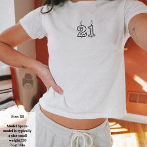 May include: White XS t-shirt with a black graphic of the number 21, styled like a birthday candle. The model's measurements are: bust 34 inches, chest 33 inches, waist 27 inches, hips 32 inches, and height 5' 4".