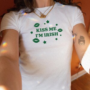 May include: White t-shirt with the text "KISS ME I'M IRISH" in green, along with green lips and star graphics. The shirt is short-sleeved and appears to be made of a soft material. The shirt is worn with blue jeans.