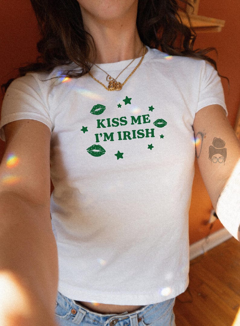 May include: White t-shirt with the text "KISS ME I'M IRISH" in green, along with green lips and star graphics. The shirt is short-sleeved and appears to be made of a soft material. The shirt is worn with blue jeans.