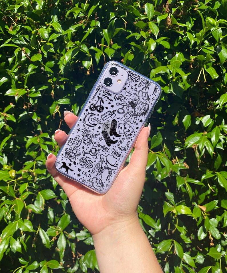 Puede incluir: Funda transparente para tel&eacute;fono con un dise&ntilde;o de tem&aacute;tica vaquera en blanco y negro. La funda presenta ilustraciones de botas de vaquero, un sombrero, herraduras y las palabras "Howdy Cowboy Tears" y "Wild".