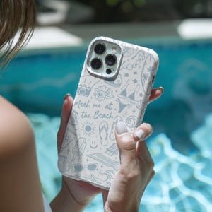 K&ouml;nnte beinhalten: Eine wei&szlig;e Handyh&uuml;lle mit Strandmotiv, mit Illustrationen einer Kamera, Sonnenbrille, Bikini und dem Text "Meet me at the Beach". Die H&uuml;lle wird in der Hand einer Person in der N&auml;he eines Pools gehalten.