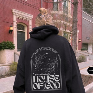Puede incluir: Sudadera con capucha negra con un estampado gr&aacute;fico blanco de un olivo con el texto "Soy como un olivo que florece en la casa de Dios".