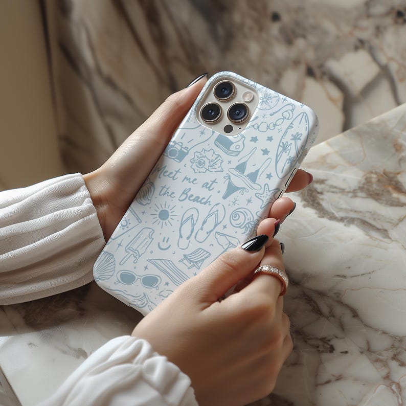 K&ouml;nnte beinhalten: Eine wei&szlig;e Handyh&uuml;lle mit einem hellblauen Strandmotiv. Die H&uuml;lle zeigt Illustrationen einer Kamera, Muscheln, Sonnenbrillen und den Text "Meet me at the Beach". Die H&uuml;lle wird in den H&auml;nden einer Person gehalten.