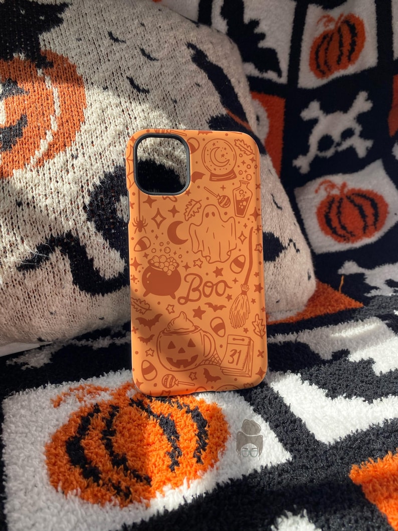 Puede incluir: Funda de tel&eacute;fono naranja con ilustraciones tem&aacute;ticas de Halloween, incluyendo un fantasma, una escoba de bruja y un caf&eacute; con leche de calabaza. La palabra "Boo" se destaca. La funda est&aacute; sobre una manta estampada con dise&ntilde;os de calabazas y calaveras.