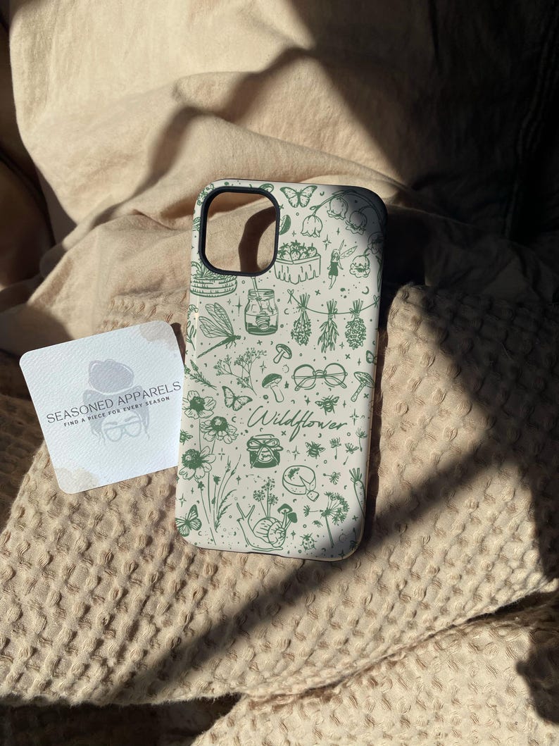 Może przedstawiać: Etui na telefon z białym tłem i zielonym wzorem polnych kwiat&oacute;w. Etui zawiera ilustracje motyli, grzyb&oacute;w, słoik&oacute;w i słowo "Wildflower". Obok znajduje się wizyt&oacute;wka z napisem "Seasoned Apparels".