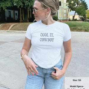 May include: White t-shirt with the text "COOL IT, COWBOY" in gray lettering. The model is wearing light blue jeans. The image also includes a size chart that says the model is wearing an XL for a baby tee fit.