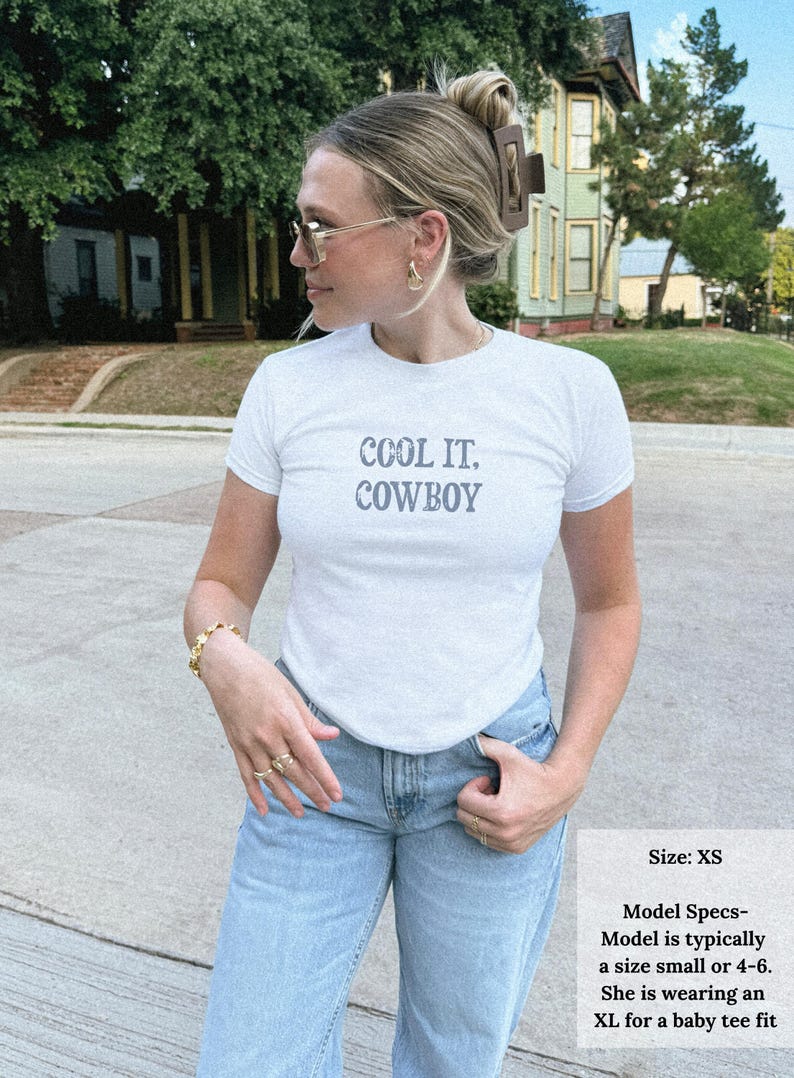 May include: White t-shirt with the text "COOL IT, COWBOY" in gray lettering. The model is wearing light blue jeans. The image also includes a size chart that says the model is wearing an XL for a baby tee fit.