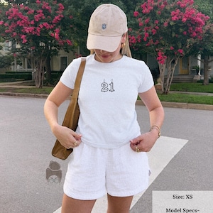 May include: White t-shirt with the number "21" in a black outline, paired with white shorts and a beige baseball cap. The outfit is completed with a brown shoulder bag. The image is taken outdoors.