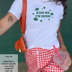 May include: White t-shirt with the text "KISS ME I'M IRISH" in green, paired with red and white checkered shorts. An orange purse hangs from a shoulder. The model specs are: size XS, weight 120 lbs, bust 34a, chest 33 in, waist 27 in, hips 32 in, height 5' 4".