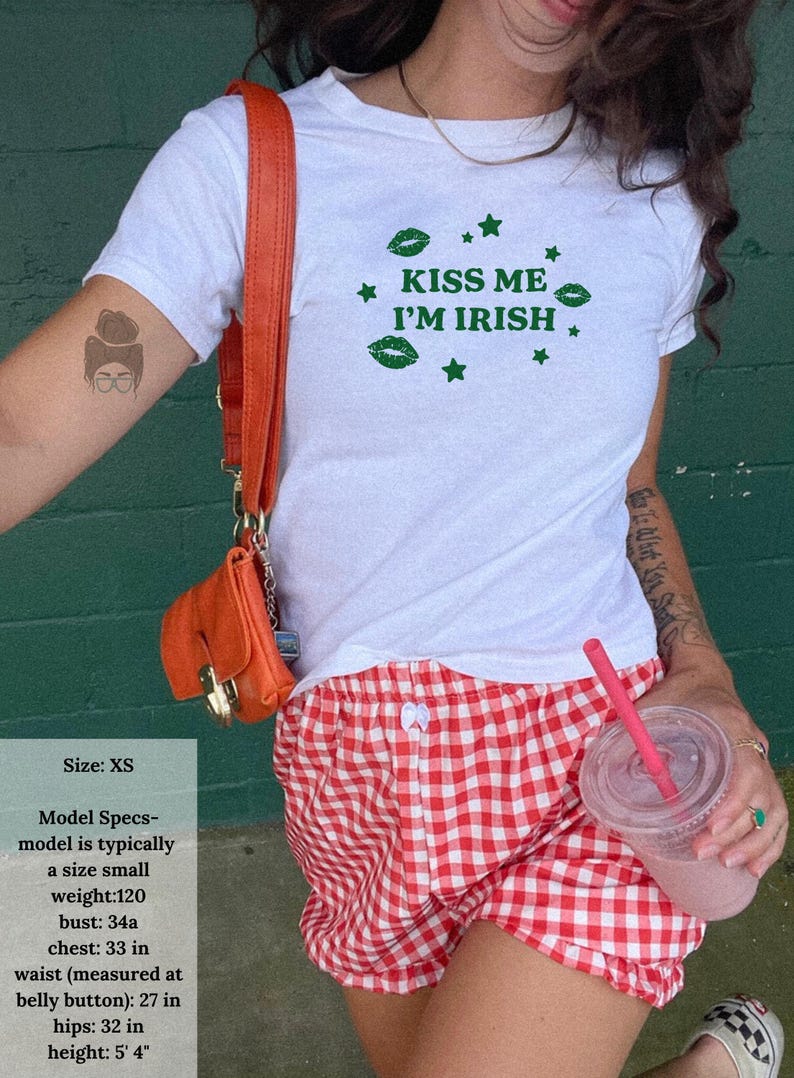 May include: White t-shirt with the text "KISS ME I'M IRISH" in green, paired with red and white checkered shorts. An orange purse hangs from a shoulder. The model specs are: size XS, weight 120 lbs, bust 34a, chest 33 in, waist 27 in, hips 32 in, height 5' 4".
