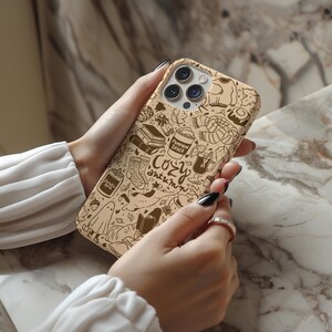May include: A tan phone case with a brown doodle design of autumn-themed illustrations. The design includes books, a pumpkin spice latte, socks, and the words "Cozy Autumn". The case is held in a person's hands.