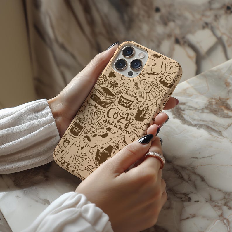 May include: A tan phone case with a brown doodle design of autumn-themed illustrations. The design includes books, a pumpkin spice latte, socks, and the words "Cozy Autumn". The case is held in a person's hands.